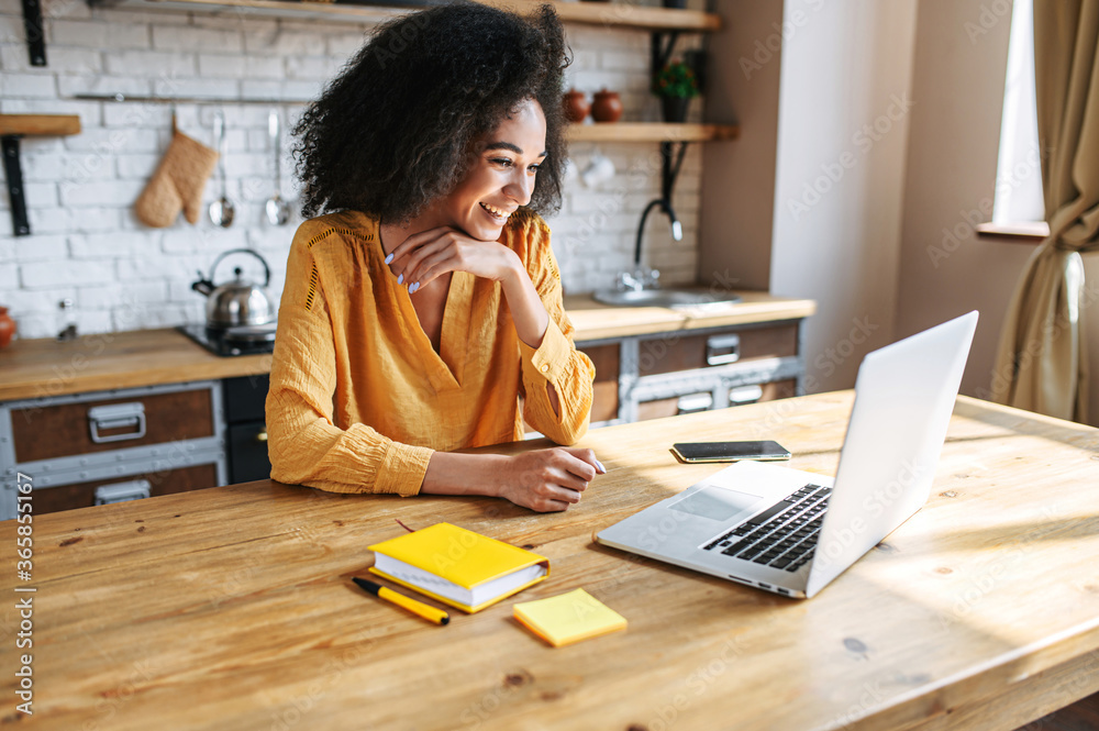 A cheerful mixed-race girl uses laptop for remote work or home leisure while sitting in the kitchen at home. Side view a nice girl with an afro hairstyle looks at screen with a smile